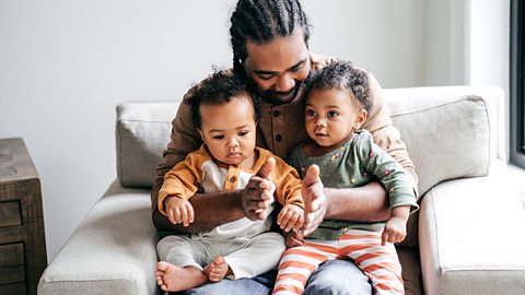 A dad sits on the sofa with 2 babies. He's clapping his hands while singing to them.