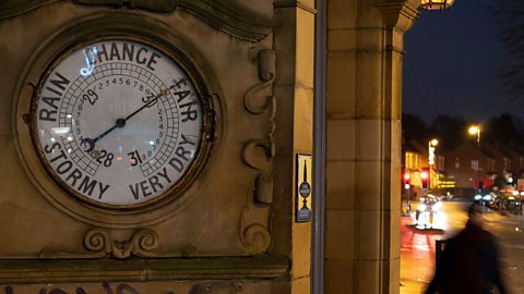 A close up of a barometer built into the a wall on the side of a building - in front of a busy road.