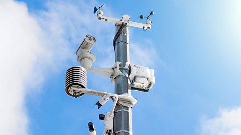 A weather station with a monitoring system and video cameras attached to it – all in front of a blue sky and clouds.