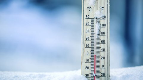 A thermometer standing up in snow, with snow stuck on it, that is showing freezing temperatures in both Celsius and Fahrenheit.