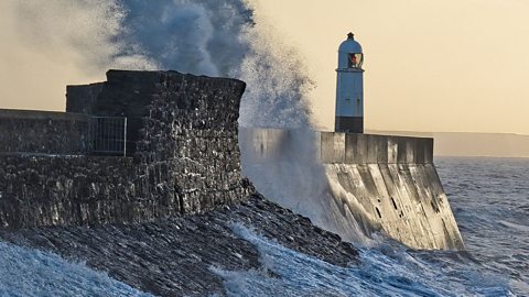 A wave crashing onto a sea wall with a lighthouse