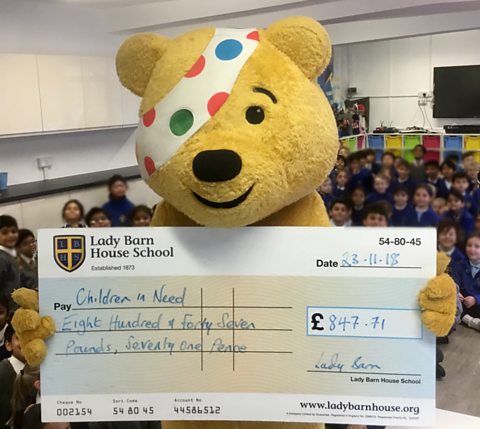 Pudsey, the yellow bear who is the Children in Need mascot, at a primary school holding an oversize cheque.