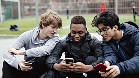 Smiling friends looking at teenage boy's phone while sitting against soccer field