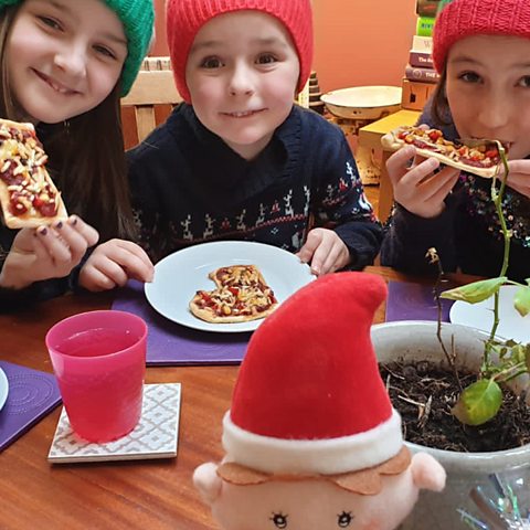 Three children are eating Christmas Pizza stockings in Christmas jumpers.