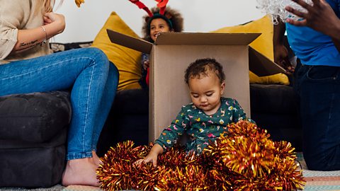 A baby sits on floor, playing with tinsel, while her family unpack the xmas decorations.