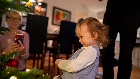 A young girl is decorating the christmas tree. Her mum is recording her on her phone.