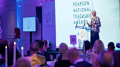 A photograph from the 2020 Teaching Awards, showing a lady giving a speech on stage, and people sitting at tables watching. 