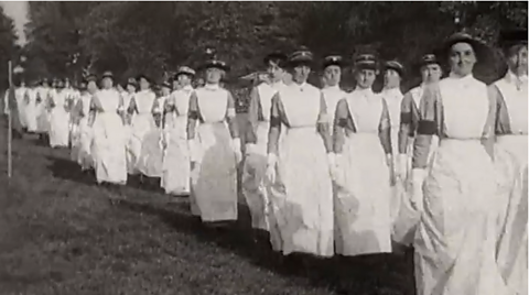 A black and white photograph of women dressed in white outfits, walking to work during the First World War.
