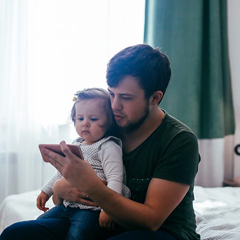 A dad and his daughter sit on a bed. They are on Facetime.