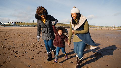 A grandparent, a mum and a daughter walk on a beach in the winter.