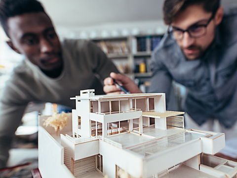 Close up shot of scale model house on table. A black architect is leaning over, looking at the model. A white architect with glasses is pointing at it with a pen. 