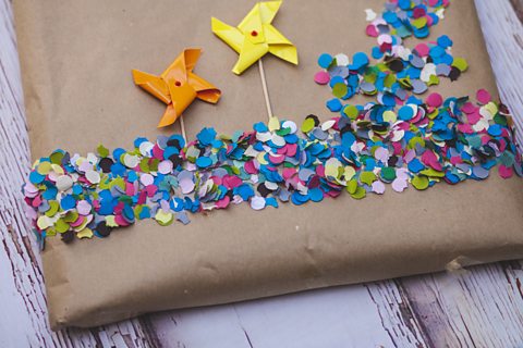 A gift wrapped n brown paper, multi-coloured confetti and hand-made paper windmills.