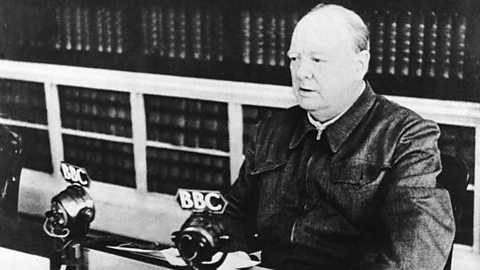 A black and white photograph of Winston Churchill sitting at a desk delivering a speech. There are two microphones on the desk in front of him.