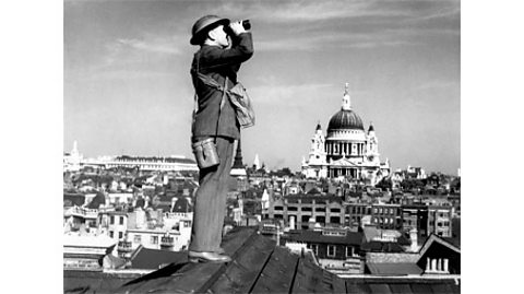 A black and white photograph of a member of the Observation Corps using binoculars to look over London.