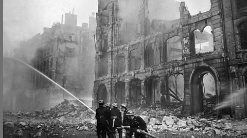 A black and white photograph of firefighters standing among ruined buildings and using a long hose to put out fires after a bombing raid in London.