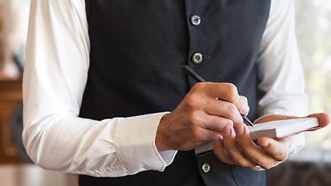 A white waiter wearing a smart black waistcoat and white long sleeved shirt. The waiter is writing on a pad.