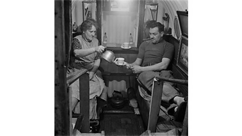 A black and white photograph of a couple sharing a cup of tea in their Anderson shelter.