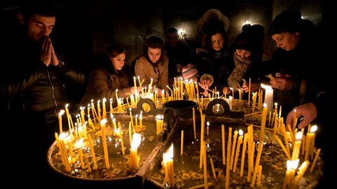 A group of worshippers pray and light candles in a darkened room, all dressed in dark clothing.