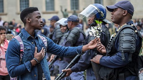 A man gives a sprig of flowers to a heavily armed policeman holding a gun wearing an ammunition belt