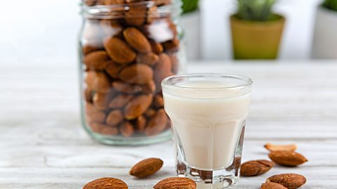 Some loose almonds and a glass of almond milk with a jar of almonds in the background.