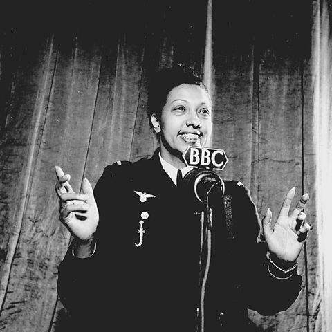 A black and white photograph of Josephine Baker. She is wearing a military style jacket with her hair in a bun, and is standing in front of a BBC microphone.