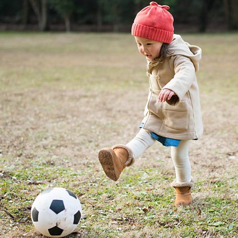 A girl in a red hat and a coat is kicking a football in a park.