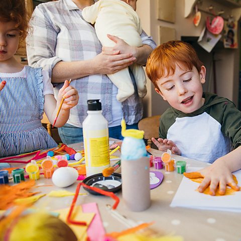 A young ginger boy and his sister play with crafts. He his finger painting. A carer stands in the background with a baby.