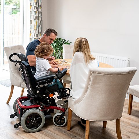 A family sits around their dining room table doing a jigsaw puzzle. There is a mum, dad and a young boy in a wheelchair.