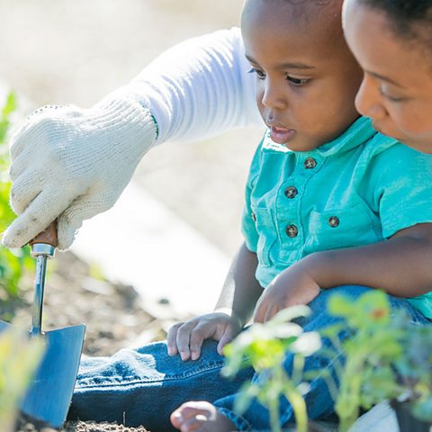 A baby boy and his mum are gardening. She is wearing gloves and holds a trowel.