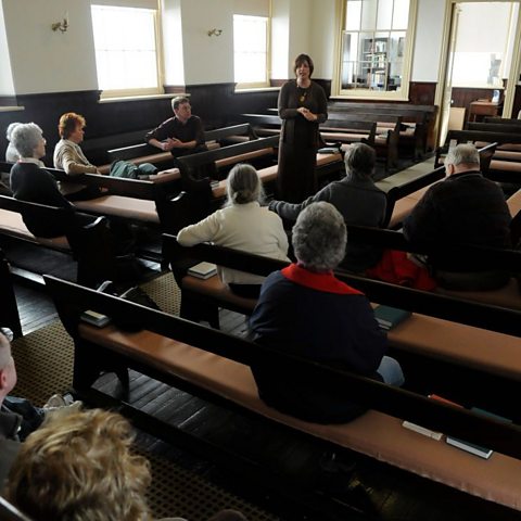 A small group of people sit on padded wooden benches in a light and airy room, with one woman stood up and addressing the others.