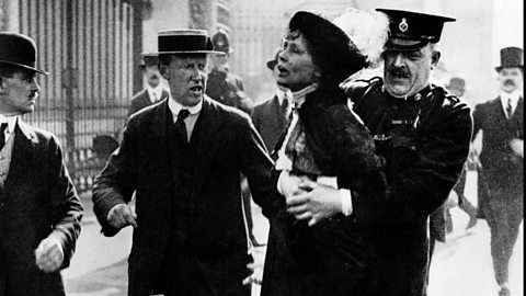 A black and white image of a woman being carted away by a police officer, with a man walking alongside looking angry.