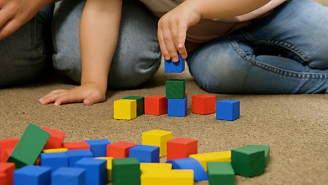 A child's hand is building a tower with multi-coloured blocks on the floor.