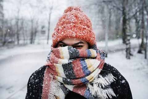 A young woman, wrapped up in cosy winter clothes, scarf and hat, stands outside while it snows.