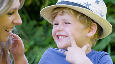 A young blonde boy in a hat is smiling at his mum and pointing to his face.