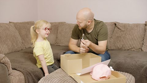 A father and his daughter sit on a sofa and play an out one out game. He is holding a spoon. There is a box on the table in front of them.