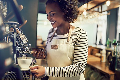 A Black waitress wearing a balck and white stripy top and a cream apron prepares a coffee using a coffee machine.