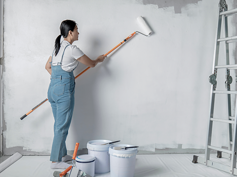 A female Asian woman wearing blue dungarees is a painting a wall white with a roller.