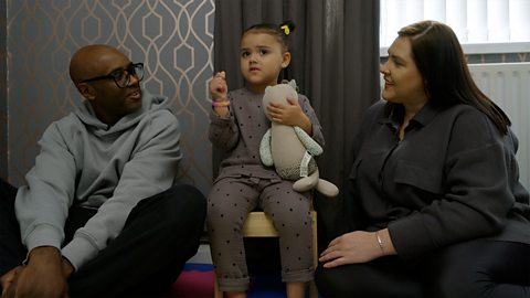 A couple and their 3-year-old child sit in their living room. Their child is holding a teddy.