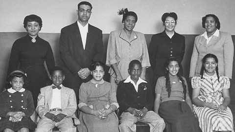 A photograph showing six children sitting on chairs and four women and one man standing behind them