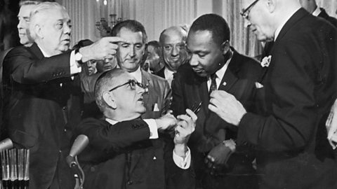 A photograph of President Lyndon B Johnson shaking hands with Dr Martin Luther King surrounded by people