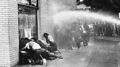 A photograph of groups of people huddled against a wall, trying to escape the water cannon being sprayed at them