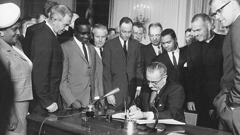 A black and white photograph of Lyndon B. Johnson signing the first Civil Rights Act, with a group of people standing around his desk.