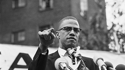 A black and white photograph of Malcolm X standing in front of some microphones to deliver a speech at a rally.