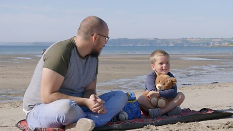 A father and his young son sit on the beach. The boy holds a teddy. They are playing the tomorrow game.