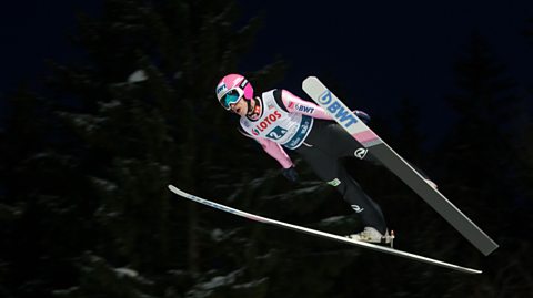 A ski jumper wearing a pink helmet and jumpsuit soars through the air, with his skis held in a V position beneath him