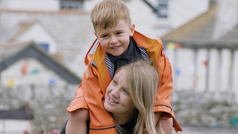 A boy in an orange coat sits on his mother's shoulders. They are playing a fun rhyming game together.