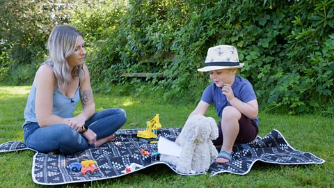 A mum and her son sit on a picnic blanket. They are joined by some toys. The boy holds crayons in his hand to share.