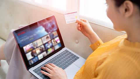 A woman holding a small card and using that to refer to whilst typing something into a video subscription streaming service.