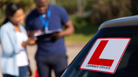 A close up of an L plate on a car with the words 'Learner Driver' under it. In the background are two Black people looking at a clipboard.