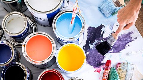 Pots of paint on a mat in three colurs. Blue. Orange and yellow. A paintbrush spreads paint on the mat that the paint sits on.